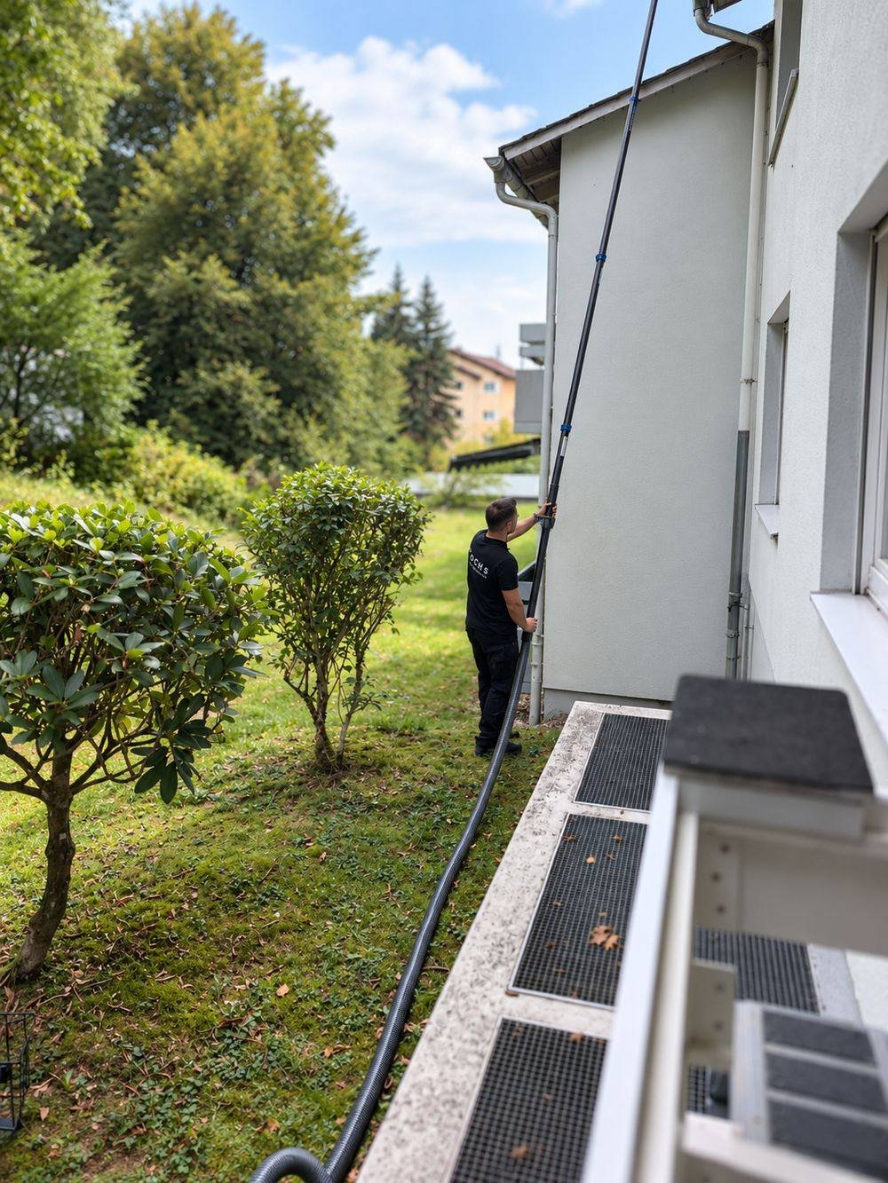Seitliche Ansicht derselben Dachrinnenreinigung: eine Person arbeitet mit dem Teleskoprohr an der Dachrinne, der Absaugschlauch verläuft an der Hauswand entlang durch eine Grünanlage.