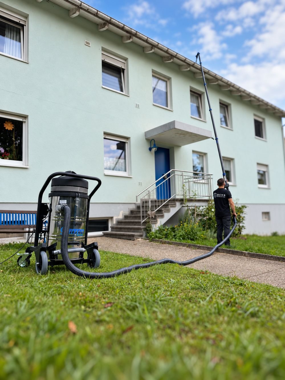 Person im OCHS-Shirt reinigt mit einem Teleskoprohr eine Dachrinne, davor das Sauggerät auf der Wiese, im Hintergrund ein Mehrfamilienhaus.